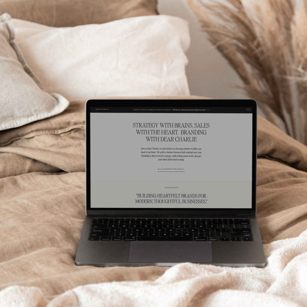 Laptop featuring Dear Charlie's website situated on a bed with neutral throws and cushions, with pampas grass in the background.  