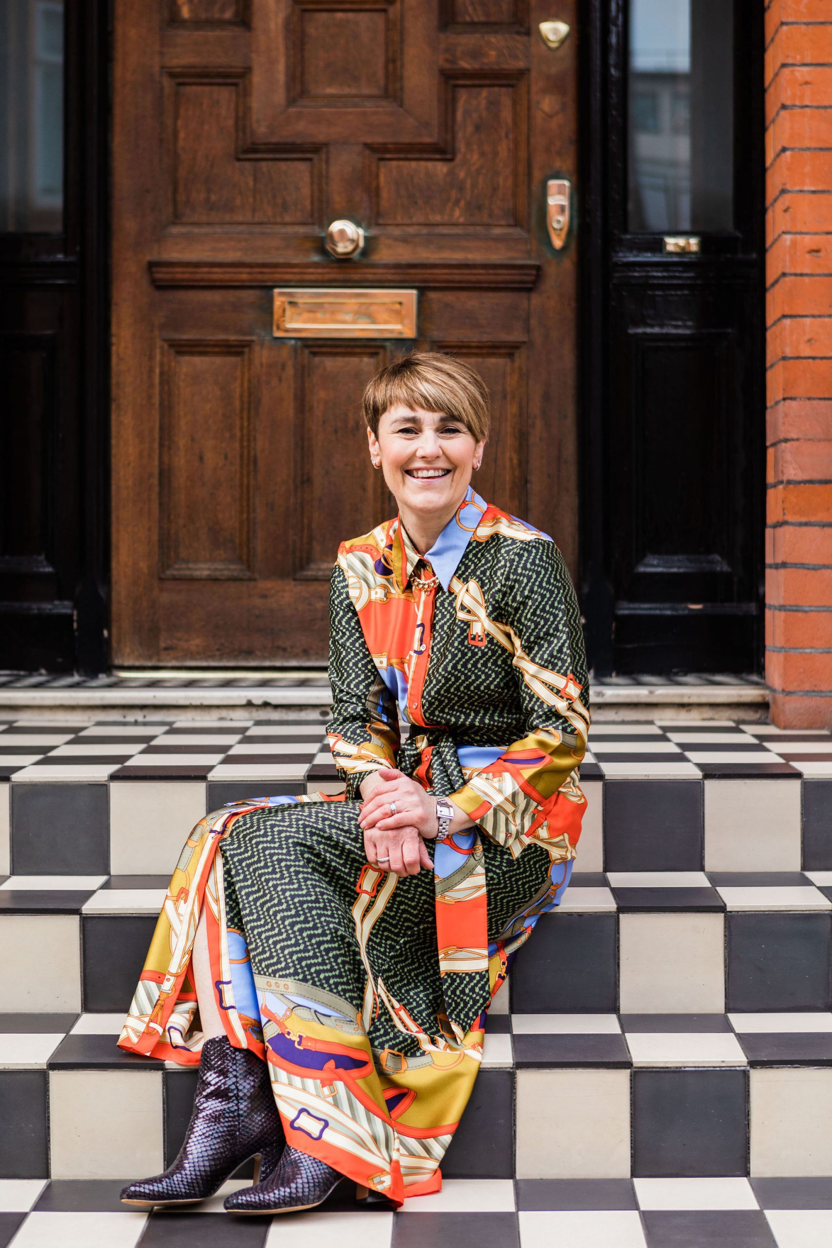 Lisa Talbot, featured on the 'Brand Essentials Podcast,' sits smiling on a checkered doorstep, dressed for success in a vibrant, patterned maxi dress and chic ankle boots, epitomizing how a wardrobe can shape personal brand and confidence in business.